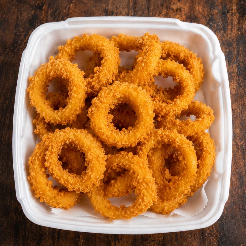 Golden-brown, crispy onion rings in a white takeout container on a dark wooden surface.