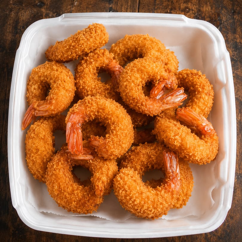 Golden-brown breaded shrimp rings and pieces in a white takeout container on a wooden surface.
