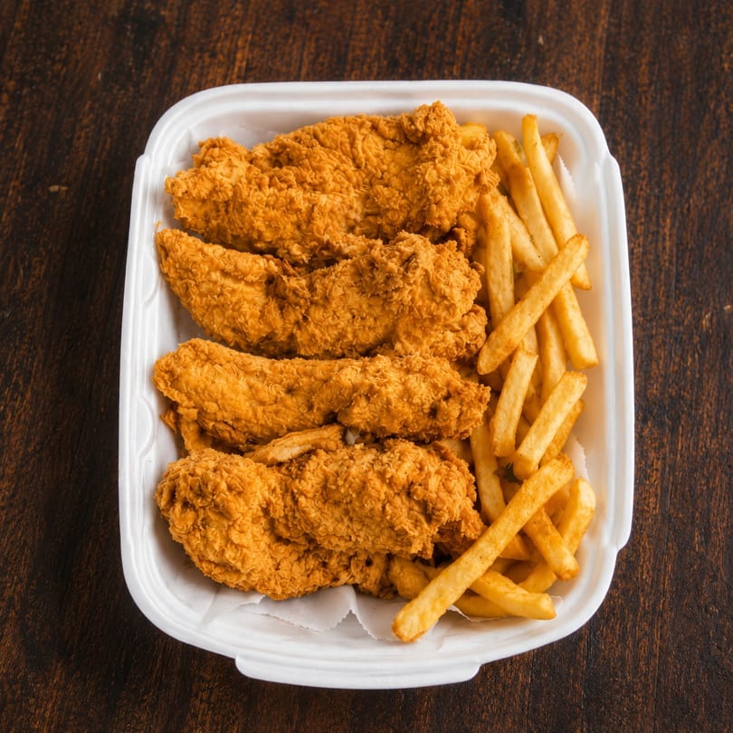 Crispy fried chicken tenders and golden french fries in a white takeout container on a wooden table.